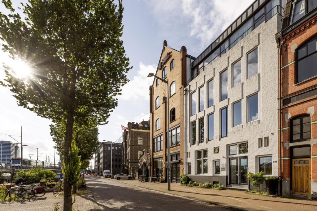 Straatbeeld van de De Ruijterkade in Amsterdam met meerdere karakteristieke kantoorgebouwen en een zonnige boomrijke stoep.